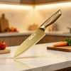 A stainless steel knife with a gold-colored handle rests on a marble surface next to vegetables.