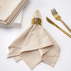 A stack of ten beige cotton table napkins tied with a gold patterned ribbon on a white surface.