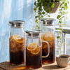Three glass water pitchers with stainless steel lids filled with iced tea sit on a woven mat near a window.