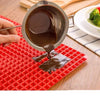 A hand pours a dark sauce from a metal pitcher onto a red silicone BBQ pizza mat.