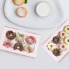A pink box contains various decorative candy rings and a jar of white cream on a white plate.