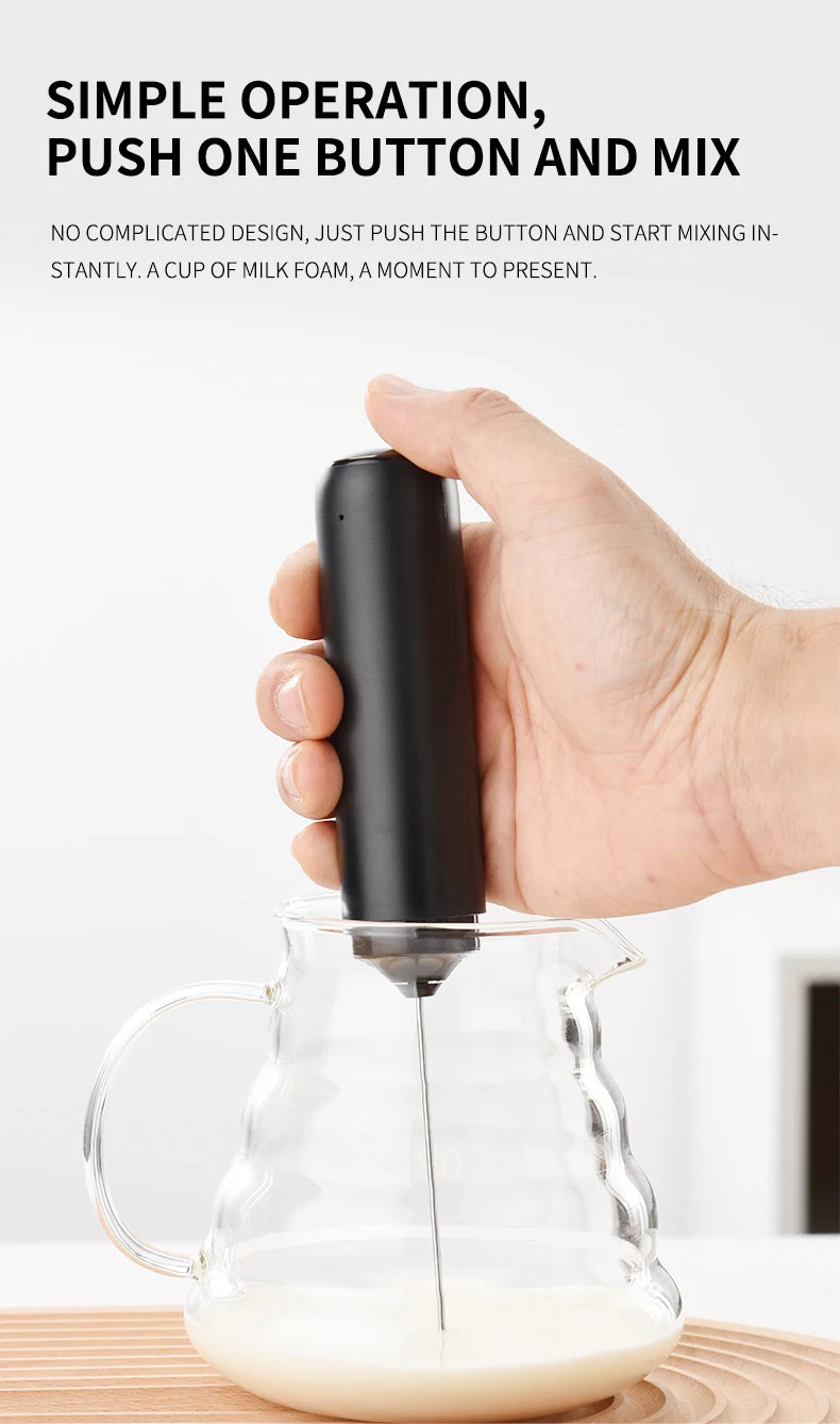 A hand is using a black handheld electric milk frother to mix milk foam in a glass pitcher.