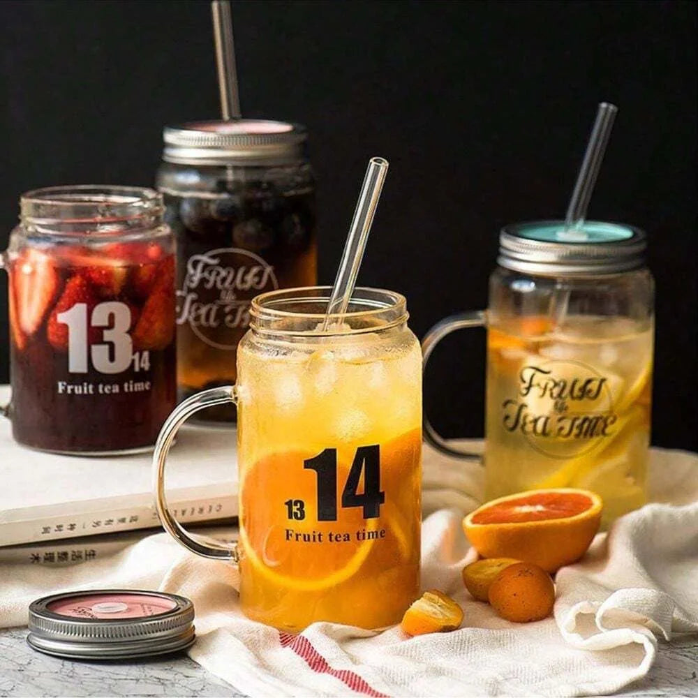 A glass jar of orange juice sits next to a glass jar of fruit tea, with reusable glass smoothie straws and a cleaning brush v