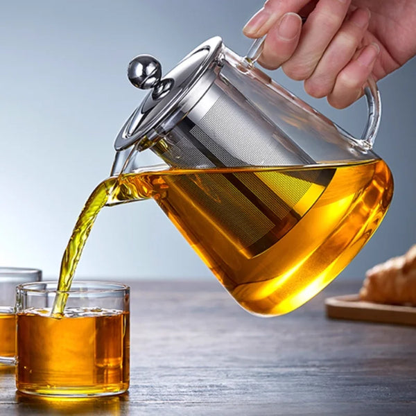 Eine Hand gießt bernsteinfarbenen Tee aus einer Glas-Teekanne mit Edelstahlfilter in eine Glastasse.