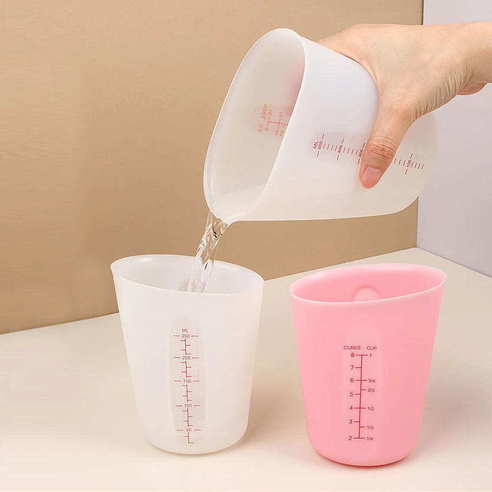 A hand pours liquid from a white silicone measuring cup into a pink silicone cup.