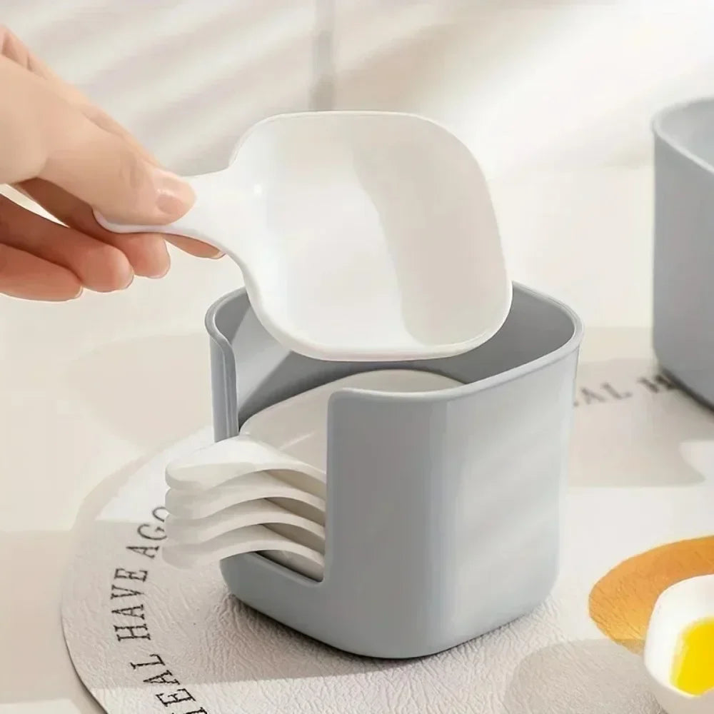 A hand is holding a white plastic sauce dish with a spoon, resting on a gray plastic dish set.