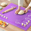A person rolls out dough on a purple silicone mat with a wooden rolling pin, surrounded by various food items.