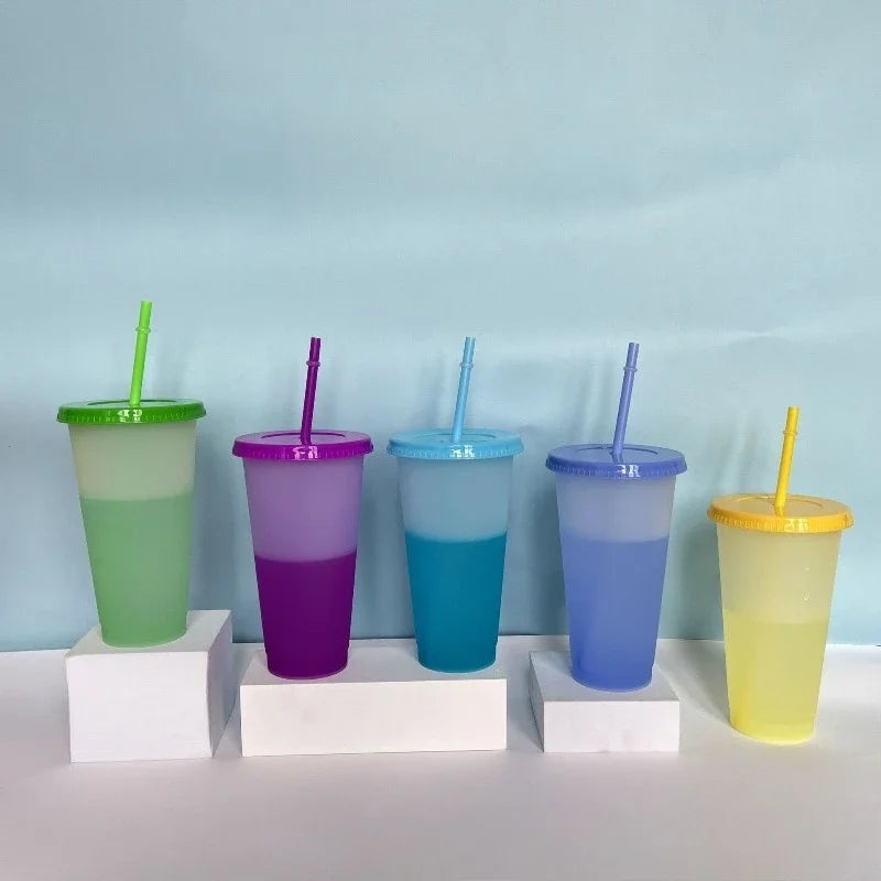 A row of ten colorful reusable tumblers in various shades of green, purple, and blue with straws set against a light blue bac