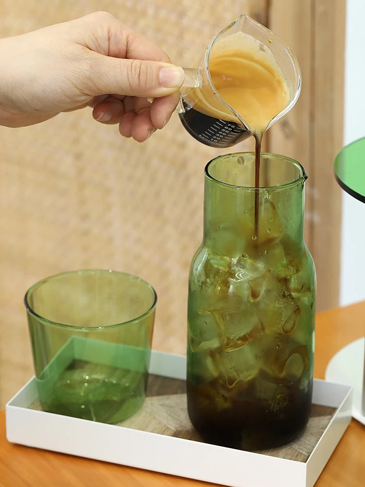 A hand pours amber liquid from a glass into a green glass mug, set on a white tray.