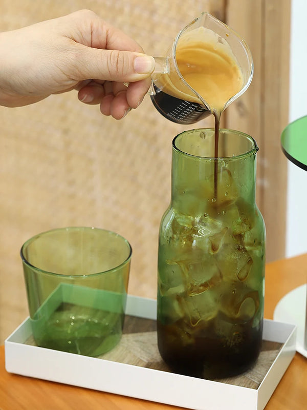 A hand pours amber liquid from a glass into a green glass mug, set on a white tray.