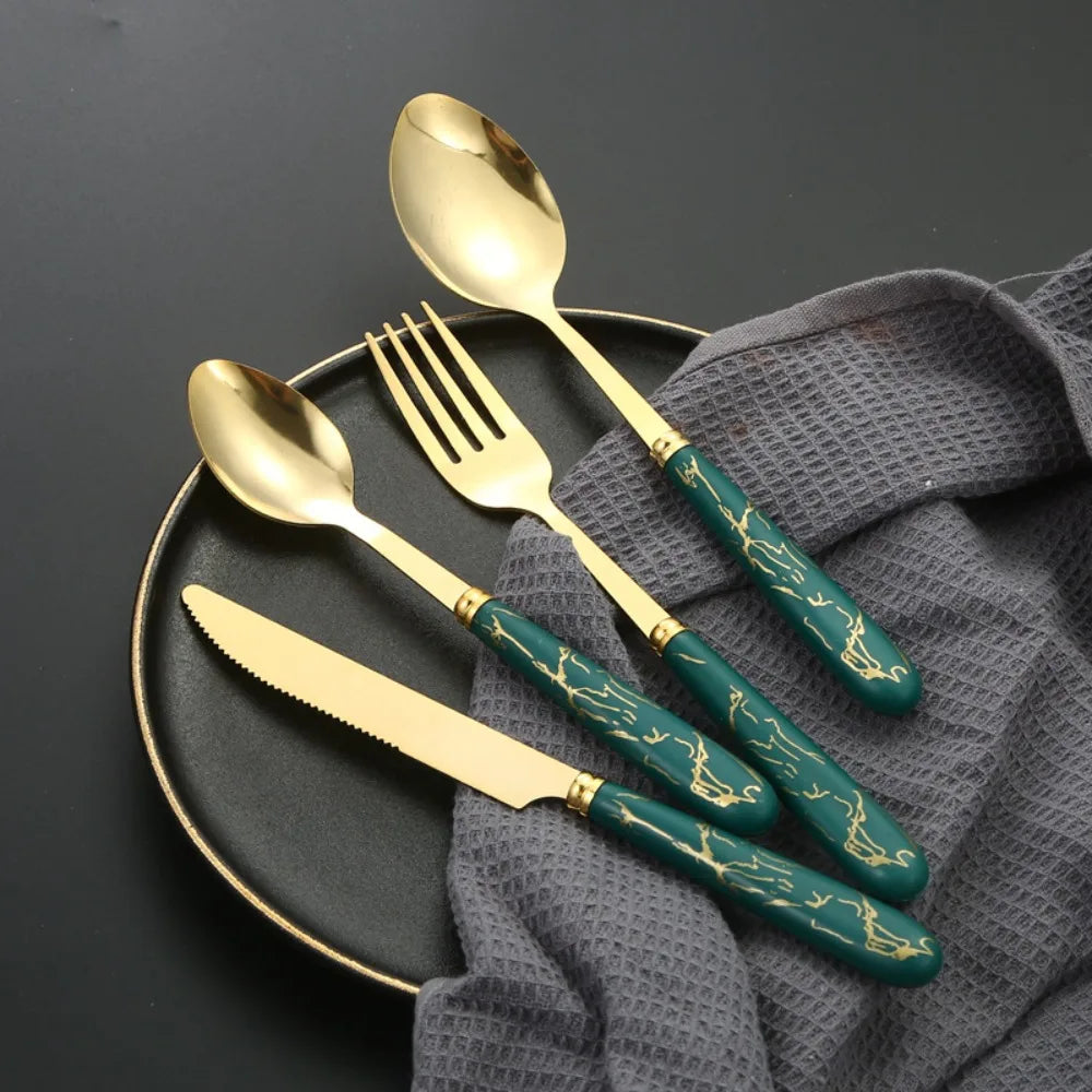 A set of gold and green marbled stainless steel cutlery including a spoon, fork, and knife displayed on a dark plate.
