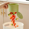 A hand is pressing a green silicone drain basket, releasing diced orange and green vegetables into a white bowl.