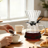 Hands prepare coffee using a manual pour over dripper and cups on a wooden table.