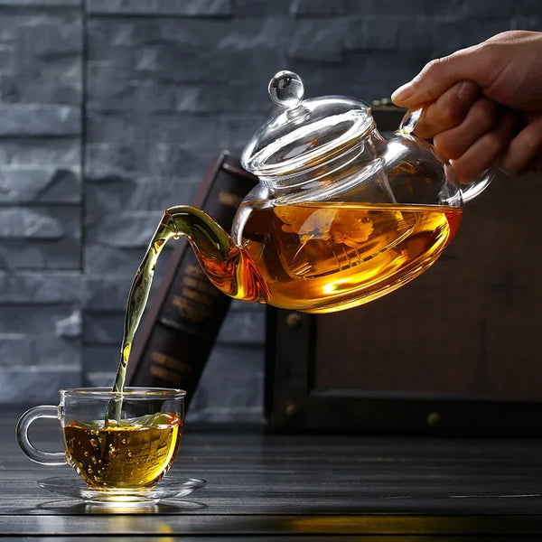 Eine Hand gießt Bernsteintee aus einer Glas-Teekanne in eine kleine Tasse vor dunklem Hintergrund.
