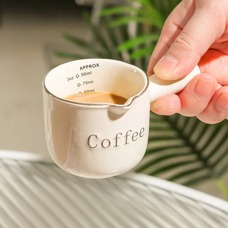 A hand is holding a white ceramic coffee cup with volume markings, showing coffee inside.
