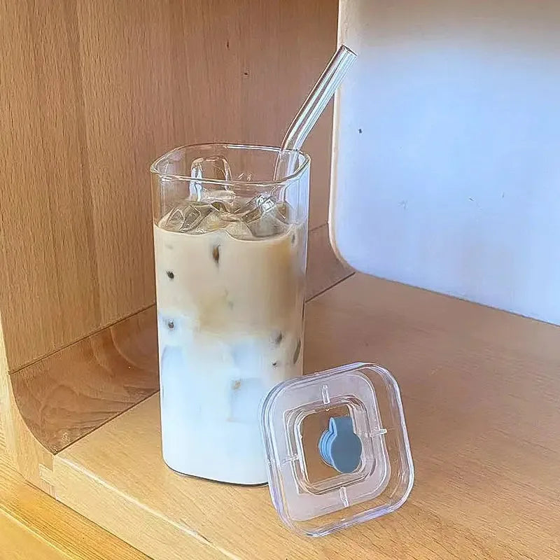 A tall glass of iced latte with foam, served with a clear plastic lid and straw on a wooden surface.