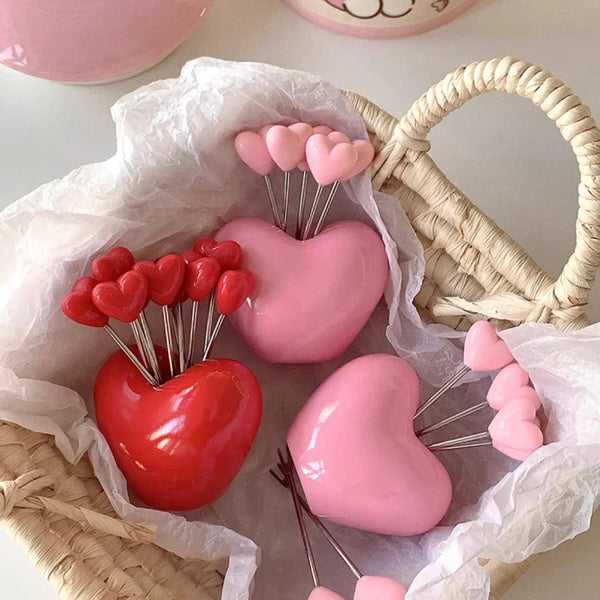 A set of stainless steel fruit and dessert forks with pink and red heart-shaped tips displayed in a white paper bag.
