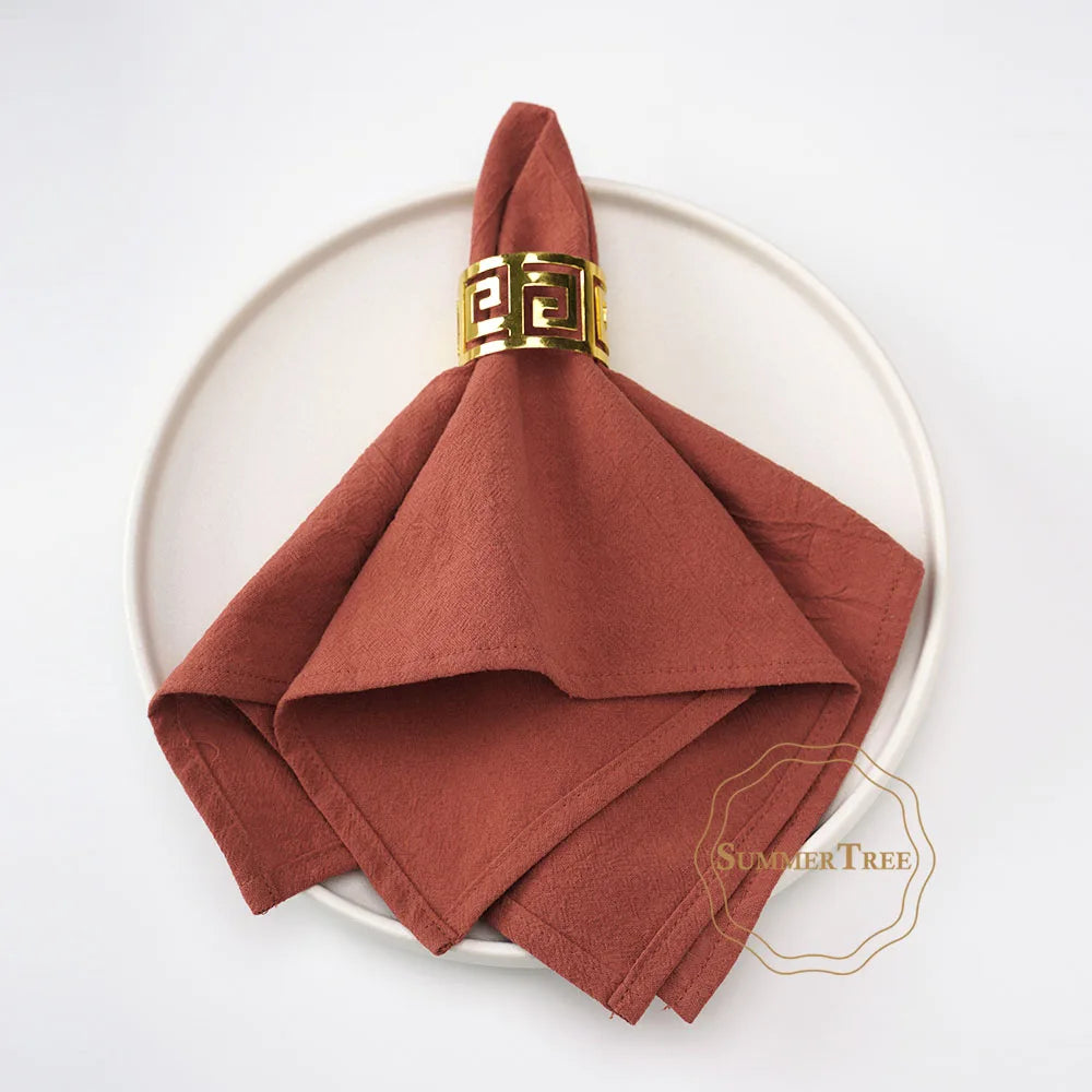 A stack of ten rust-colored cotton table napkins tied with a gold patterned ribbon, displayed on a white plate.