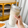 A hand holds a clear drinking glass with a lid and straw containing a light brown beverage.