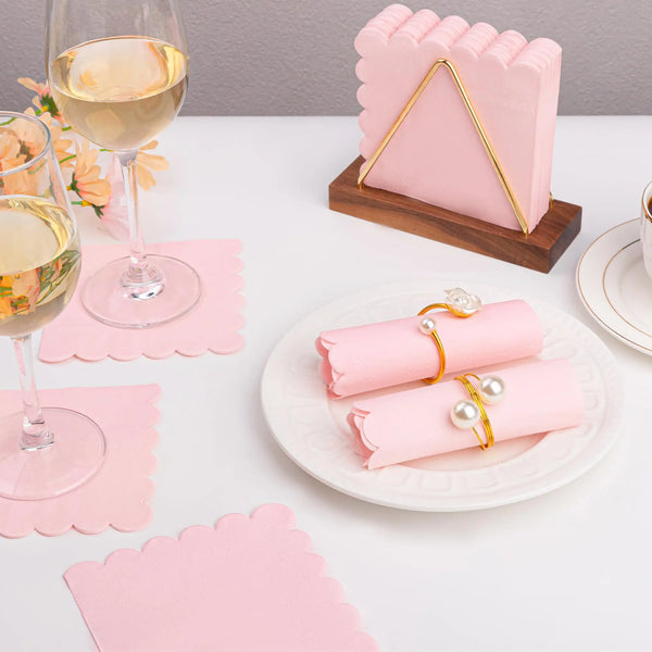 Pink scalloped edge dessert napkins displayed on a white table with a glass of white wine and a pink gift box.