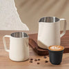 A white stainless steel coffee frothing pitcher and a black cup are displayed on a wooden surface.