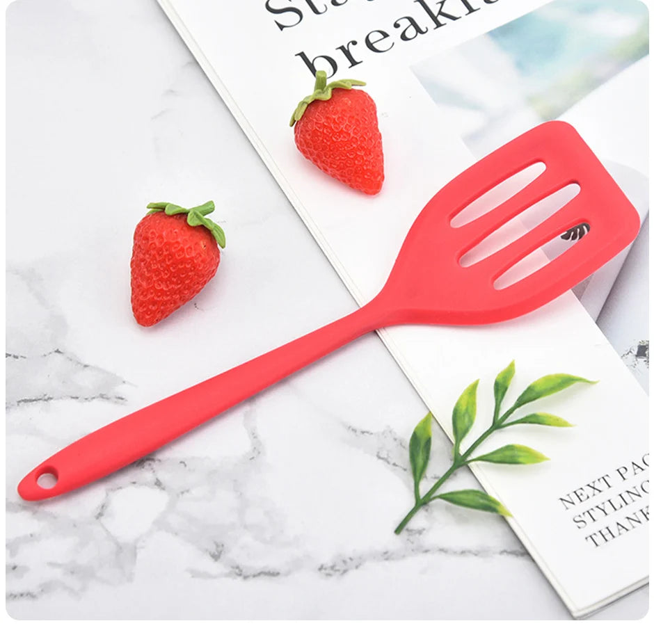 A bright red silicone flat bottom frying spatula is shown next to a fresh strawberry on a white marble surface.