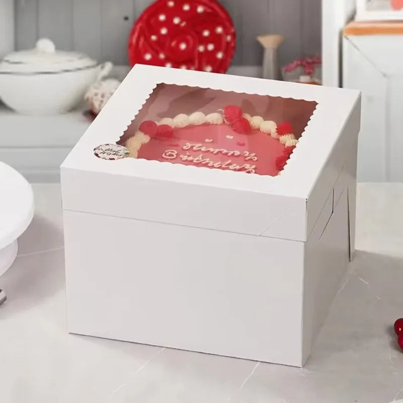 A white square disposable cake box with a clear window displaying a strawberry and blueberry decoration.