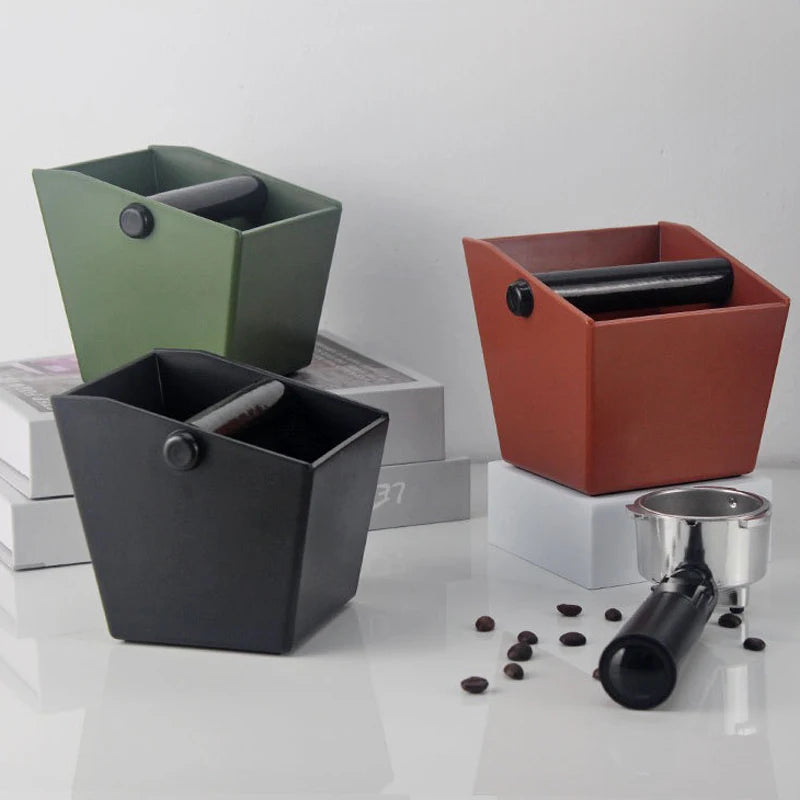 Three espresso knock boxes in olive green, black, and terracotta sitting on a white surface.