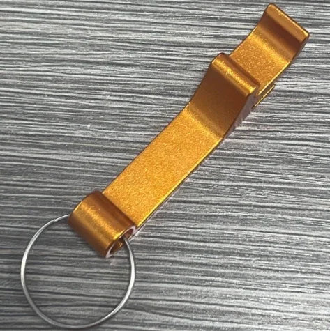 A portable metal bottle opener keychain in a golden-bronze color resting on a gray wooden surface.