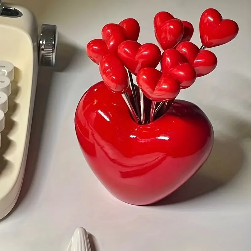 A bright red heart-shaped bowl filled with numerous small, shiny red fruit forks.