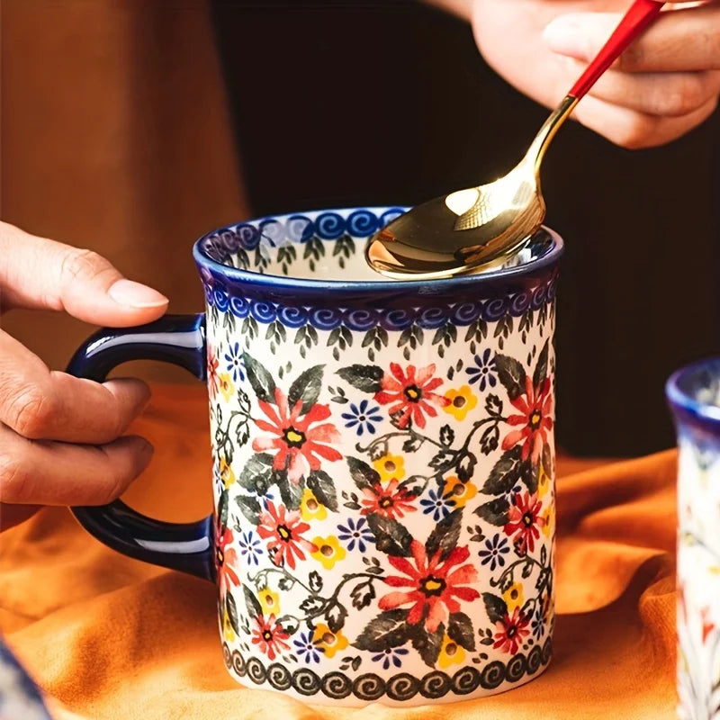 A hand holds a ceramic mug with a colorful floral pattern, and a golden spoon is dipping into the mug.