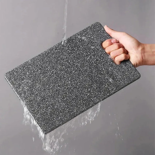 Eine Hand hält ein dunkelgraues Marmor-Anti-Rutsch-Cuttingboard unter fließendem Wasser.