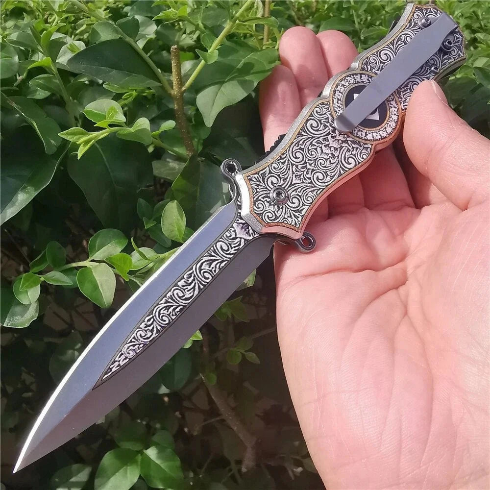 A hand holds a decorative folding utility blade knife with an ornate handle and silver blade, set against a green leafy backg