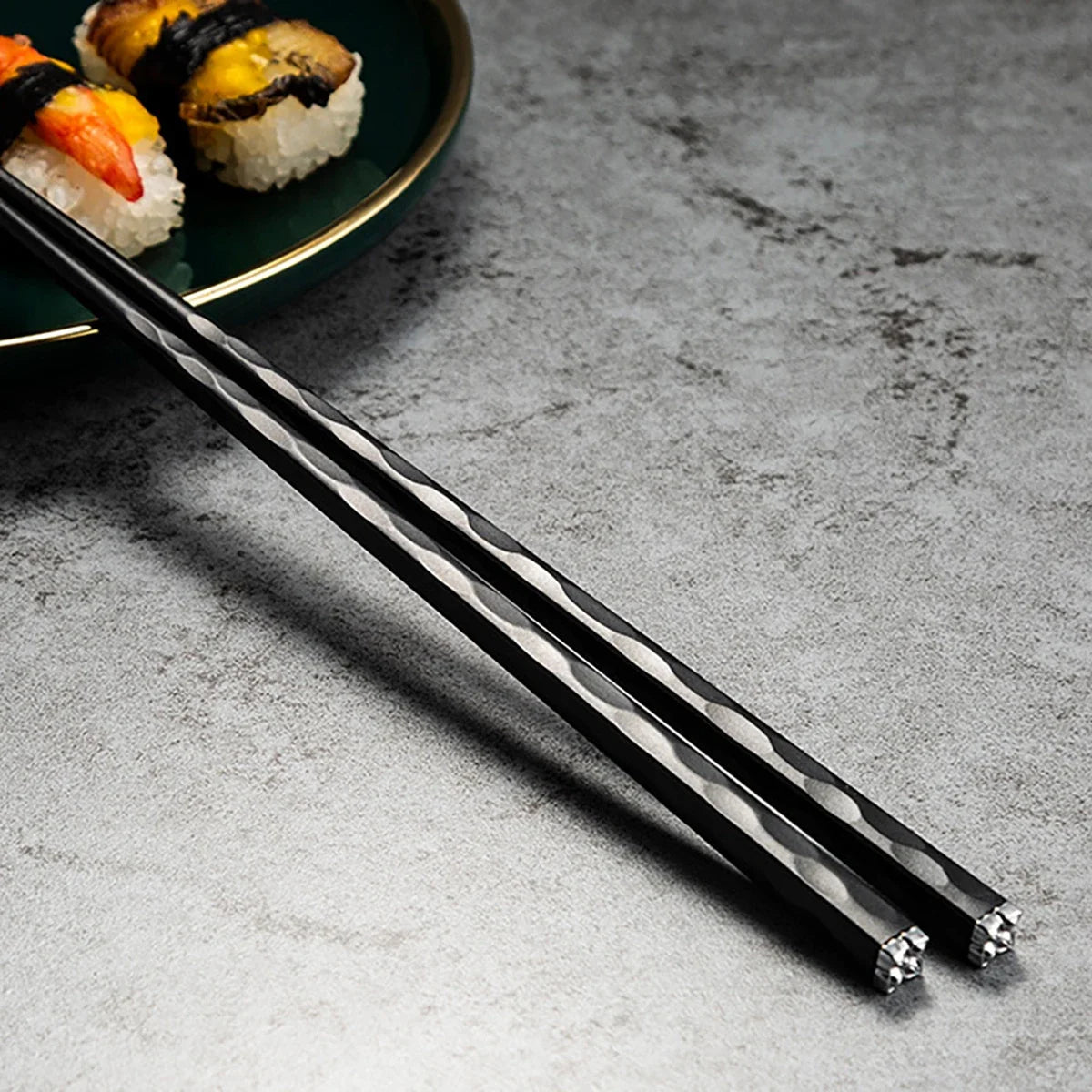 Un par de palillos reutilizables de aleación negra y plateada sobre una superficie gris junto a sushi.