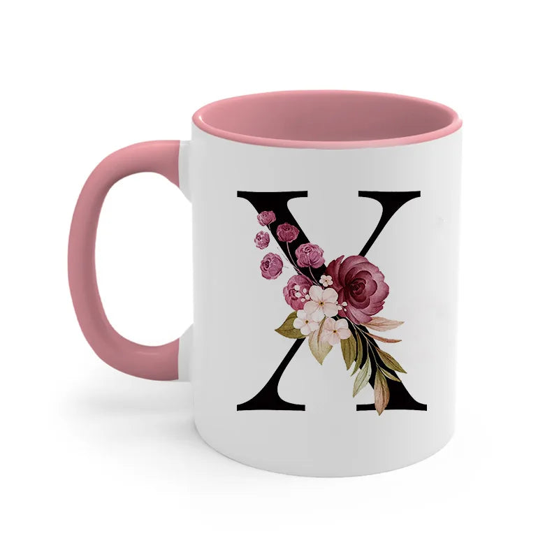 A pink ceramic coffee mug with a black stylized X monogram featuring floral accents.
