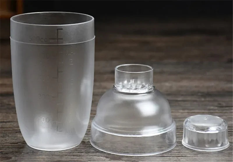 A clear plastic cocktail shaker set showing a measuring cup, a base, and a lid on a wooden surface.