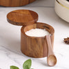 A wooden salt and spice jar with a lid and a wooden spoon filled with coarse white salt on a white marble surface.