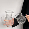 A hand holds a clear striped borosilicate glass milk pitcher with an ergonomic handle against a light background.