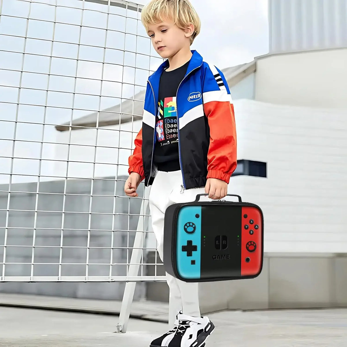 A young boy wearing a blue and red jacket stands in front of a metal fence holding an insulated thermal lunch bag.