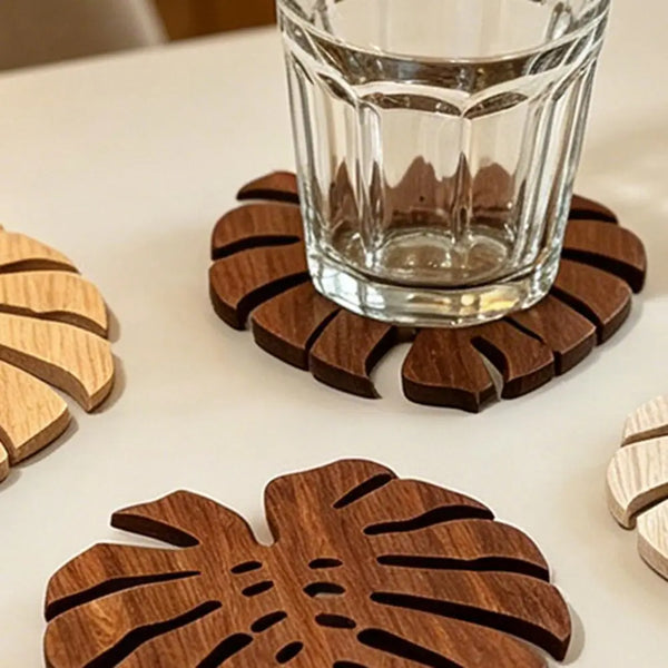 Ein Set aus fünf dunkelbraunen Holz-Tropikalblättern als Untersetzer auf einer weißen Oberfläche neben einem Glas.