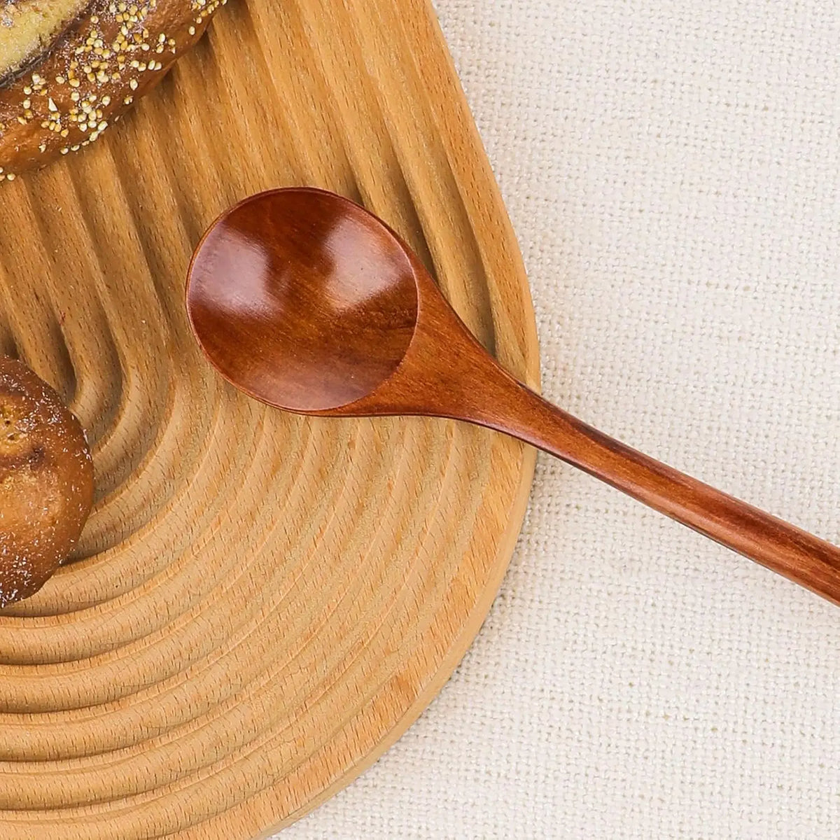A wooden serving spoon resting on a light wooden cutting board next to a loaf of bread.