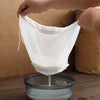 A hand is holding a white reusable fine mesh pour filter bag over a glass bowl.