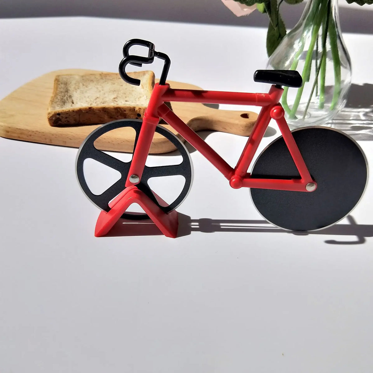 Un cortador de pizza en forma de bicicleta roja descansa sobre una tabla de madera junto a un jarrón de vidrio.
