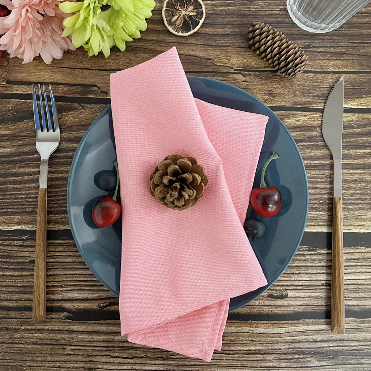 A dark blue plate holds a pink napkin and a pinecone on a rustic wooden table with cutlery and greenery.