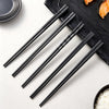 A set of five pairs of black reusable anti-slip plastic chopsticks laid diagonally on a dark slate board.