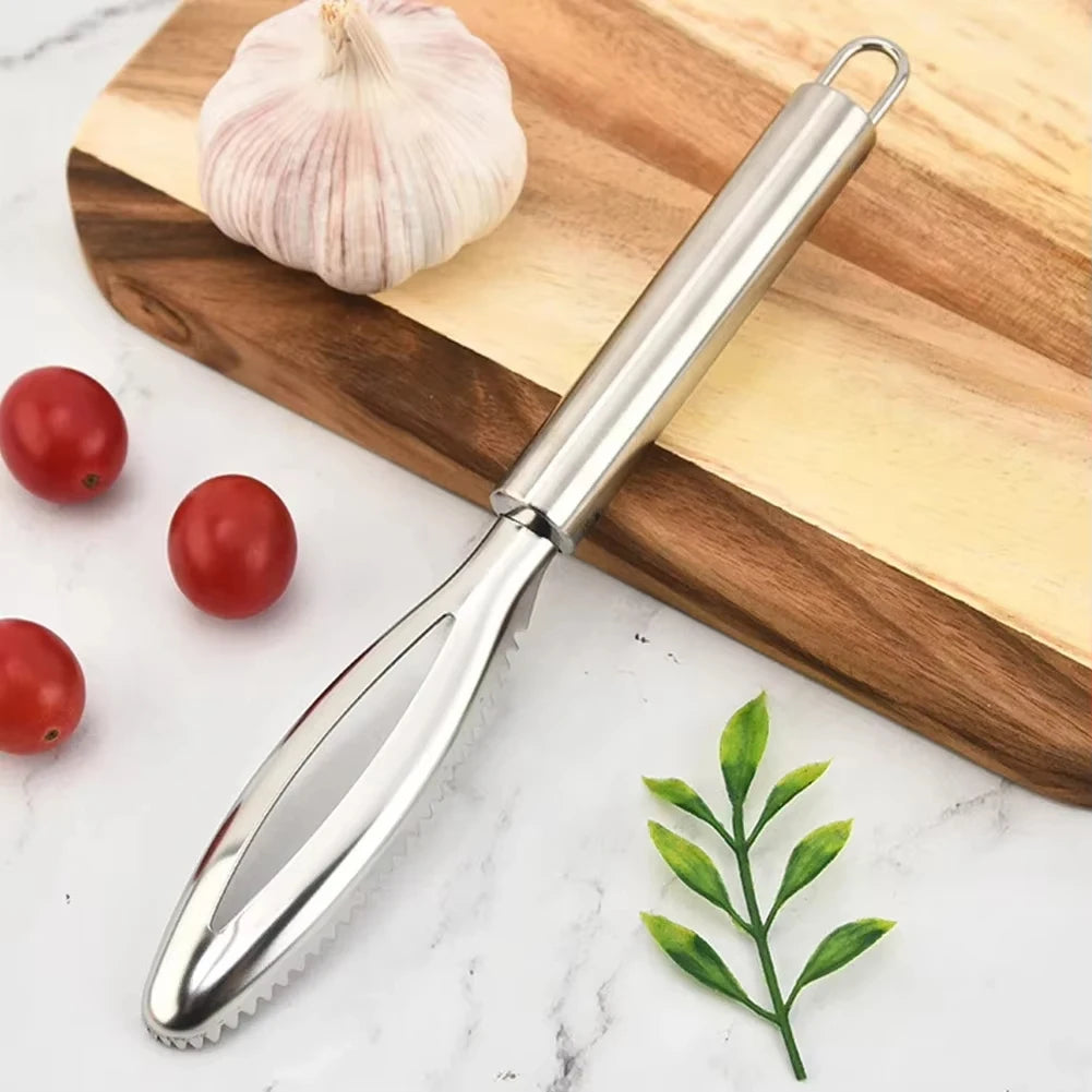 A stainless steel fish scale remover and peeler set rests on a wooden cutting board next to a garlic bulb and a sprig of gree