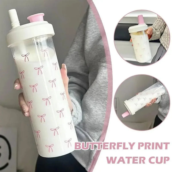 A clear water cup with a butterfly print and pink bow straw is held by a person against a gray background.