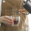 A person pours coffee from a black pour-over device into a glass mug while wearing an apron.