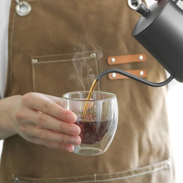 Eine Person gießt Kaffee aus einem schwarzen Pour-Over-Gerät in eine Glas Tasse, während sie eine Schürze trägt.
