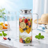 A clear glass water jug with a metal lid filled with various fruits and water on a marble surface.